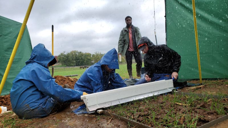 Three researchers seated on the ground and one standing collect samples from a rainfall simulator.