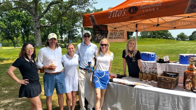 A group of golfers stand next to a bar featuring Tito's Vodka.