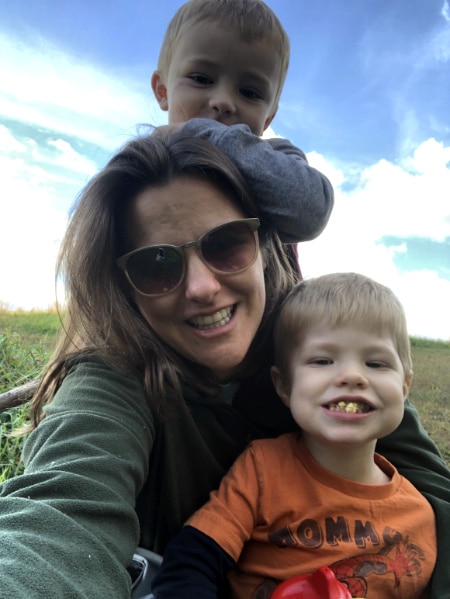Betsy Kerlin takes a selfie with her two young sons against a grassy hill and blue sky background.