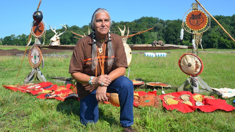 Chief Quiet Thunder kneeling outdoors in front of a display of Lenni Lenape cultural items.