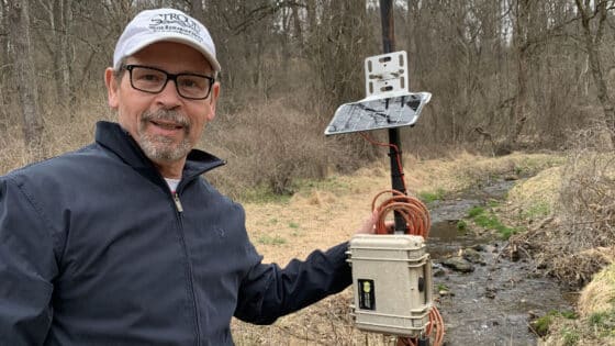 A community science volunteers stands next to an EnviroDIY Monitoring Station on the banks of a small stream.