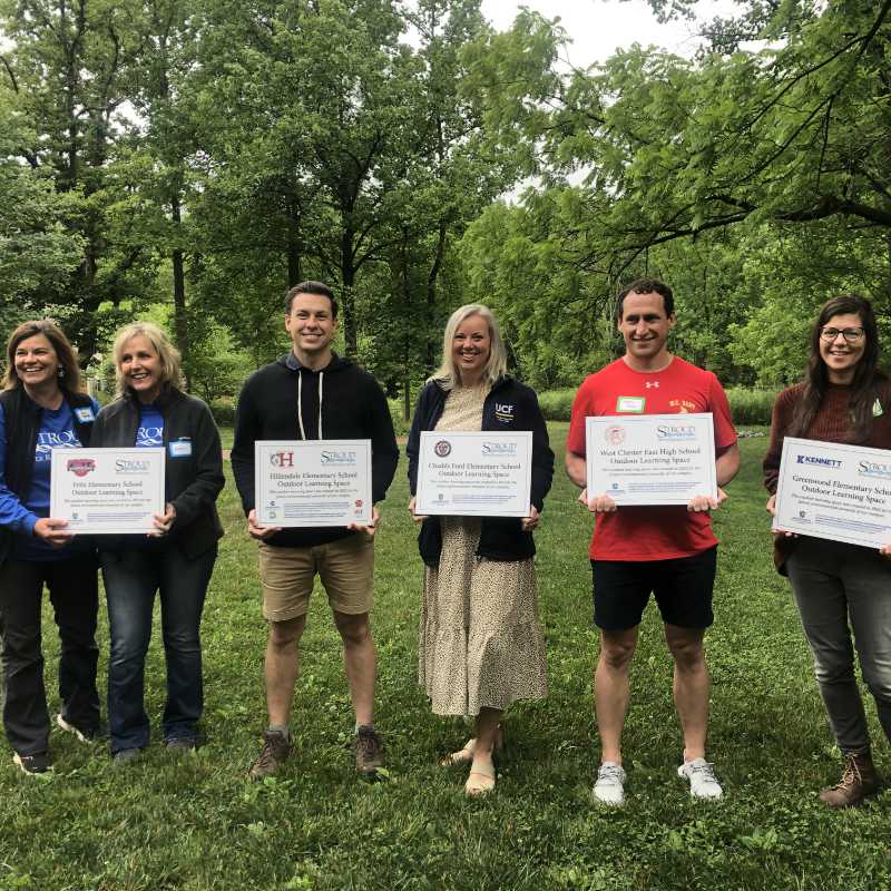 Six smiling teachers hold certificates earned at an outdoor learning workshop.