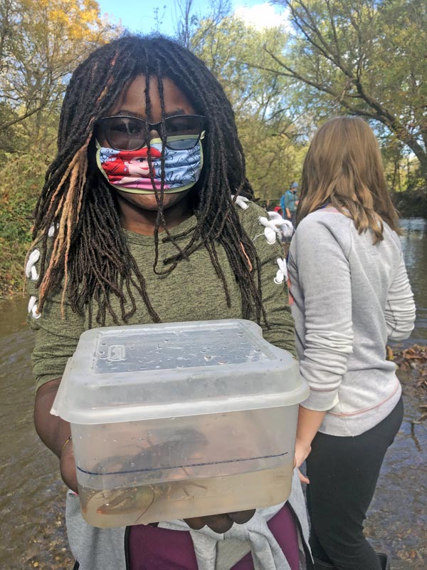 An elementary school student wearing a face mask and holding a plastic container with a crayfish inside.