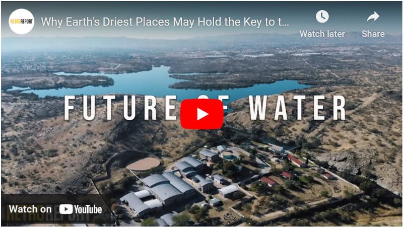 An video still showing an aerial view of buildings near a lake in an arid environment.
