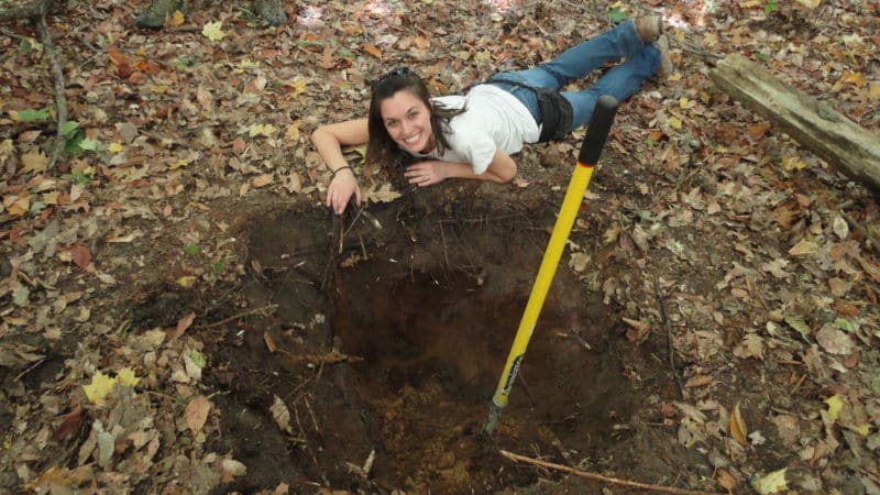 A woman smiles as she lays next to a soil pit excavated on a college field trip.