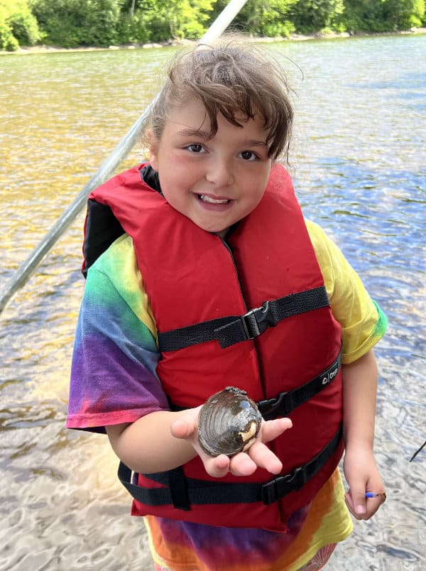 A girl wearing a red life preserver holds a freshwater mussel.
