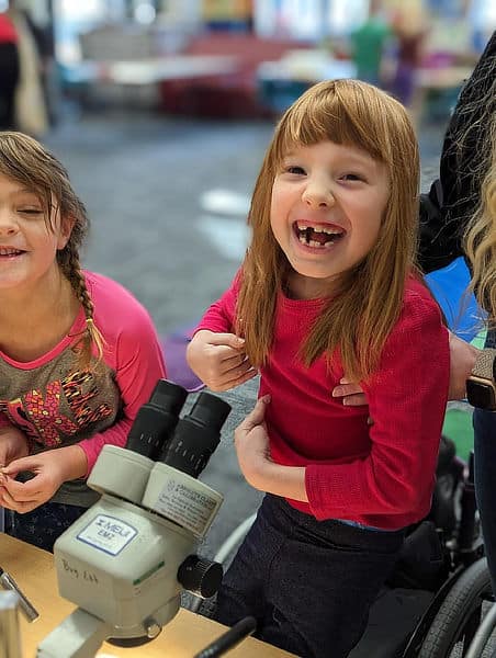A girl in a wheelchair smiles with delight at seeing creek critters under a microscope.