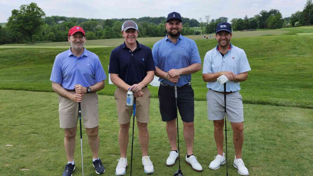 Four smiling golfers stand on a golf green holding golf clubs.