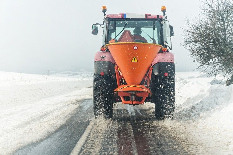 A large red vehicle spreads salt on a road surrounded by snowy fields.