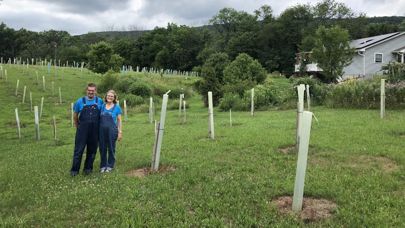 A farm couple wearing overalls smile in front of their newly planted riparian buffer.