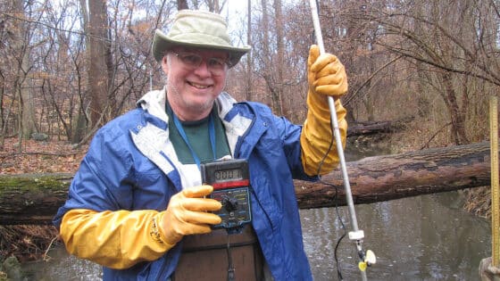 A man stands in a stream while holding water quality monitoring equipment.