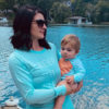 A women and an infant wearing sun protection shirts standing next to a lake.