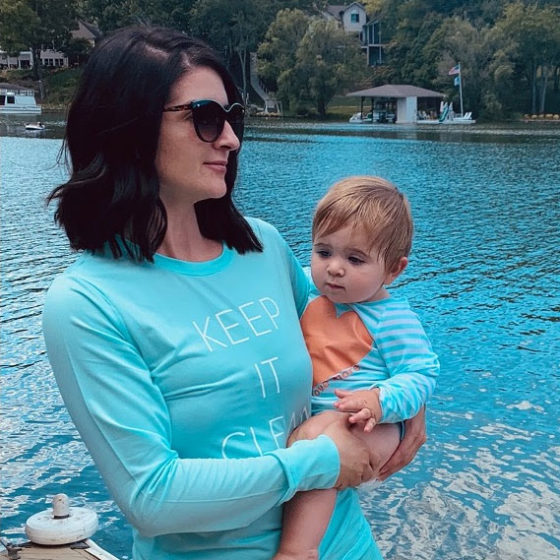 A women and an infant wearing sun protection shirts standing next to a lake.