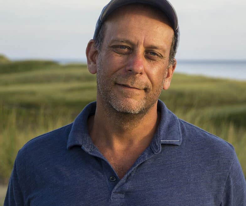 Paul Greenberg wearing a blue shirt and baseball cap with an ocean shoreline in the background.