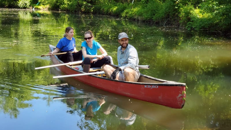 Three scientists in a red canoe on river preparing to sample milldam sediments.