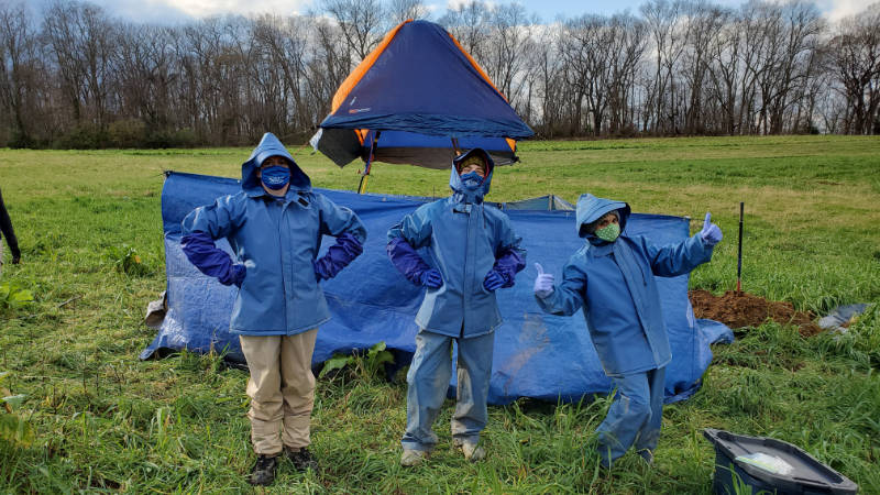 Three scientists pose in front of a rainfall simulator protected from the wind in a farm field.