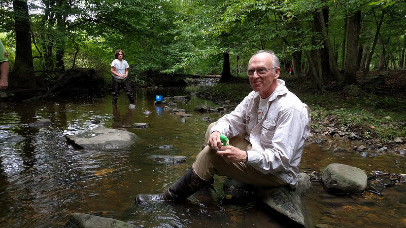 A man sits on a rock in a stream and prepares to a take a water chemistry sample.