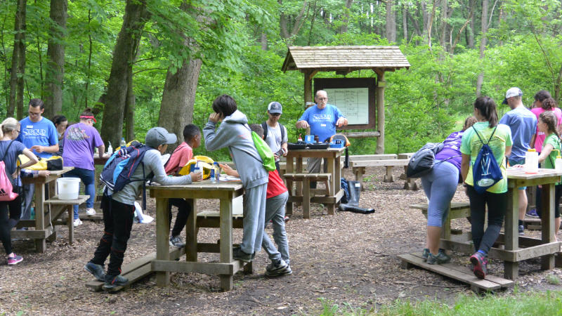 A teacher speaking to middle school students grouped around tables in an outdoor classroom.