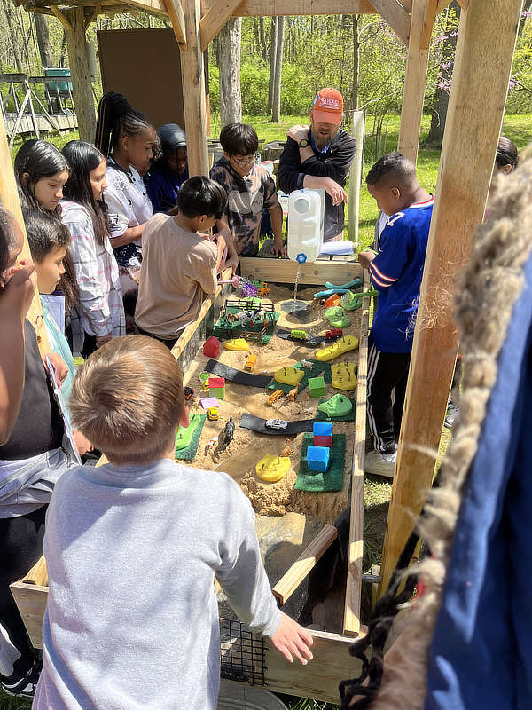 Students watch as an educator pours water into a watershed model in an outdoor classroom.