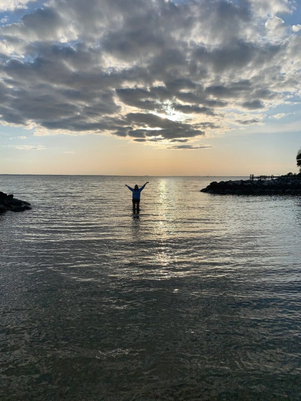 Sylvie Randall standing in a bay at sunset, with her arms raised to the clouds.