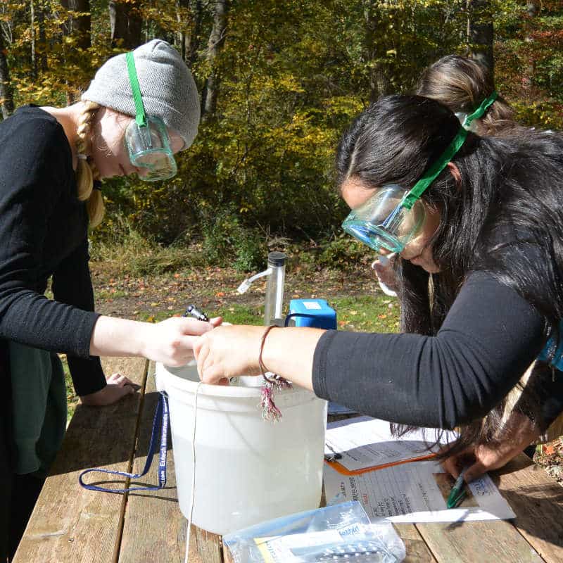 Three middle school students wearing safety googles perform water chemistry tests in an outdoor classroom.