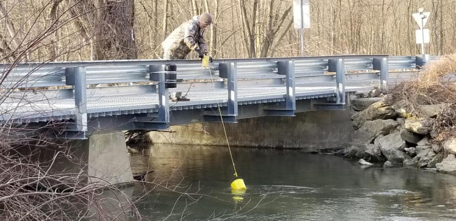 A watershed steward on a low metal bridge samples stream water using a bucket attached to a rope.