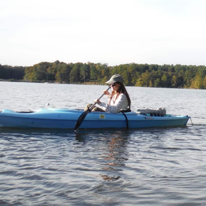 A woman in a blue kayak paddles on a lake with woods in the background.