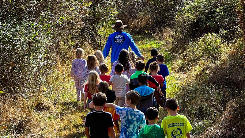 A teacher leads a group of small children along a woodland trail.