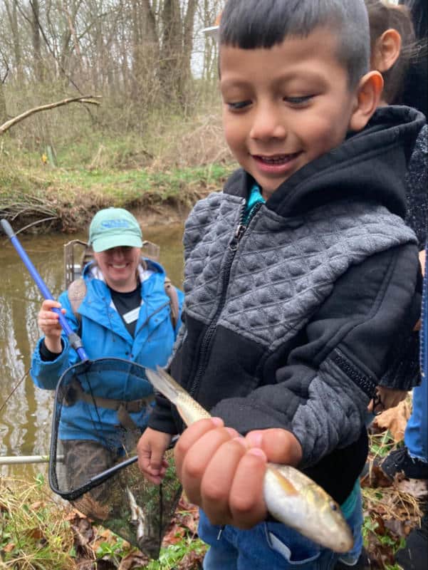 A boy stands next to a stream holding a live fish caught by a watershed research technician.